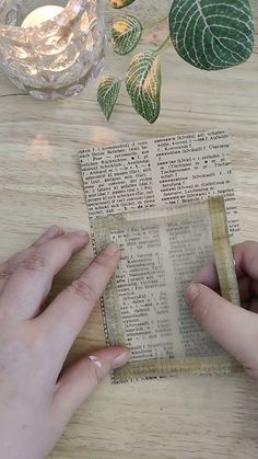 two hands holding an open book on top of a wooden table next to a candle