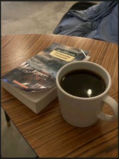 a cup of coffee sitting on top of a wooden table next to a paperback book