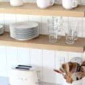 a cat sitting on top of a wooden shelf next to plates and cups in a kitchen