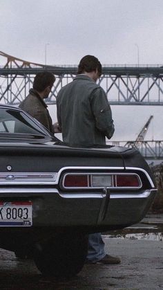 two men standing next to an old car in front of a bridge on a cloudy day