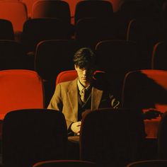 a man in a suit and tie sitting in an empty auditorium with red seats behind him