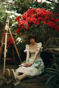 a woman sitting on the ground in front of red flowers next to a easel