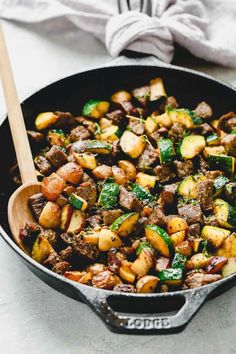 a skillet filled with meat and vegetables on top of a counter next to a wooden spoon