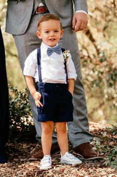 a little boy wearing a bow tie and suspenders standing next to his dad in the woods