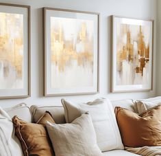 three paintings hang on the wall above a white couch with gold pillows and throw pillows