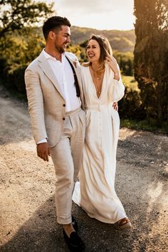 a man and woman walking down a dirt road together, dressed in beige suits with white shirts and ties