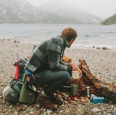 a man sitting on the ground next to a campfire with food and camping gear