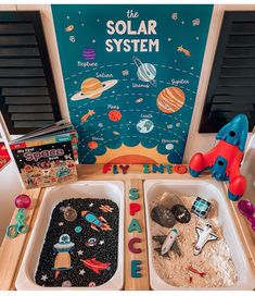 two trays filled with sand and toys on top of a wooden table in front of a solar system sign