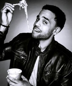 a man is eating noodles from a bowl and holding a cup in his hand while wearing a leather jacket