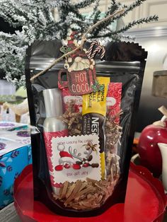 a gift bag filled with snacks and candy sits on top of a red plate in front of a christmas tree