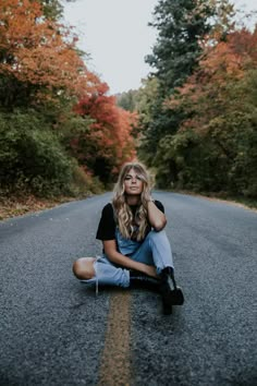 a woman sitting on the side of a road in front of trees with orange leaves