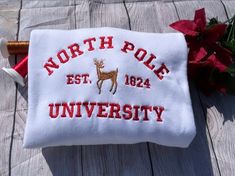 Get into the festive spirit with this North Pole University Christmas Themed Crewneck Sweatshirt. The cozy sweatshirt is perfect for anyone who want to stay comfortable while still showing off their holiday cheer. The design features a fun and colorful Christmas theme that is sure to make you stand out during any holiday event. Crafted from high-quality materials, this sweatshirt is both durable and comfortable. The crewneck style makes it easy to wear with any outfit, while the breathable fabri Embroidered Christmas Holiday Sweatshirt, Christmas Holiday Embroidered Sweatshirt, White Letter Embroidery Sweater For Winter, Christmas Cotton Sweatshirt With Embroidered Logo, Winter College Sweater With Embroidered Graphics, Winter Sweatshirt With Embroidered Logo, College Winter Sweater With Embroidered Graphics, Winter Crew Neck Sweater With Embroidered Text, Winter White Sweatshirt With Embroidered Text