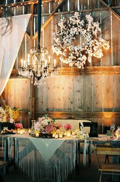 a table set up with flowers and candles in front of a wooden barn wall filled with hanging chandeliers