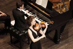 a woman playing the violin in front of a piano and two men sitting at chairs