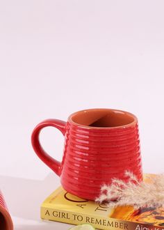 a red coffee mug sitting on top of a book next to a cup with a feather