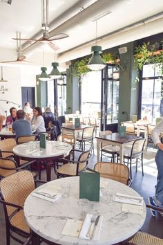 people sitting at tables in a restaurant with large windows and plants hanging from the ceiling