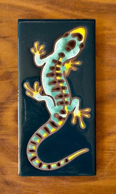 a blue and yellow lizard sitting on top of a black tile wall mounted to a wooden table