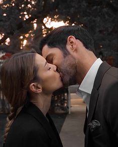 a man and woman kissing each other in front of a tree with the sun behind them