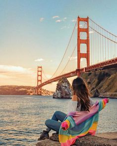 a woman sitting on top of a rock next to the ocean near a large bridge