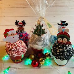 three christmas cupcakes in plastic bags on a table with lights and decorations around them