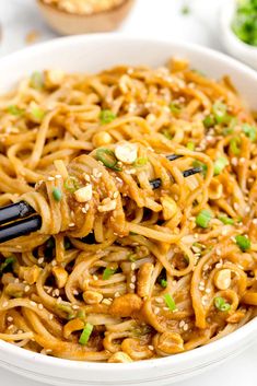 a white bowl filled with noodles and meat topped with green onions, sesame seeds and scallions