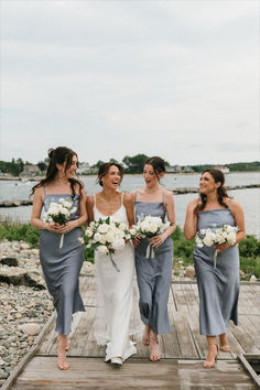 four bridesmaids laughing together on a dock by the water with bouquets in their hands