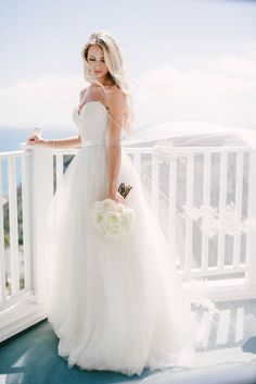 a woman in a white wedding dress standing on a balcony holding a bouquet and looking at the camera