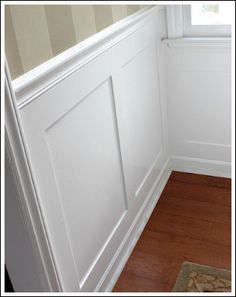 the corner of a room with white paneling and wood flooring, next to a window