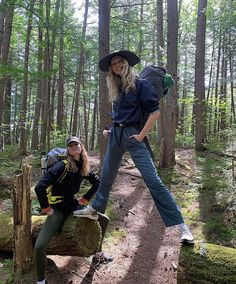 two people standing in the woods with backpacks on their back and one person sitting on a log