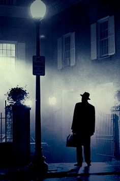 a man standing in front of a street light on a foggy night with a hat and coat