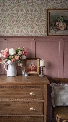 a vase with flowers on top of a wooden dresser next to a chair and wallpaper