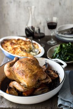 a roasted chicken in a pot with other dishes and wine glasses on the table behind it