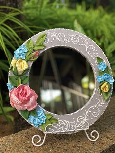 a decorative mirror with flowers painted on the front and sides, sitting on a rock