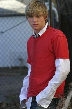 a young man in a red shirt and jeans standing next to a fence with his hands in his pockets