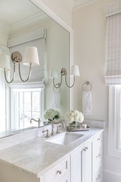 a white bathroom with double sinks and two lamps on the wall above it, along with windows