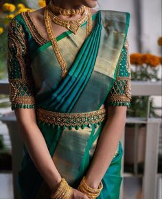 a woman in a green and gold sari with jewelry on her neck, standing next to flowers