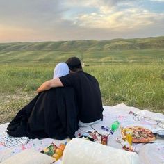 two people sitting on a blanket in the middle of a field with pizza and snacks