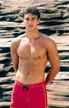 a man with no shirt on standing in front of the water wearing red swim trunks