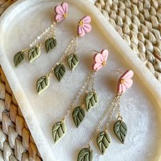 six earrings with pink and green leaves are on a white tray next to a wicker basket