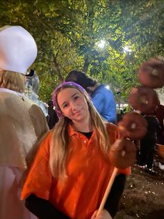 a girl in an orange shirt is holding a stick and smiling at the camera with other people behind her