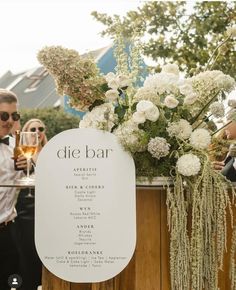 a wooden table topped with white flowers next to a tall vase filled with greenery