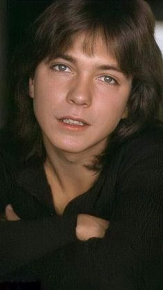 a young man with long hair and blue eyes posing for the camera, wearing a black sweater