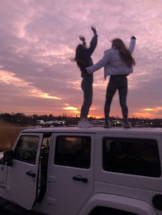 two girls standing on top of a jeep in front of the sun setting behind them