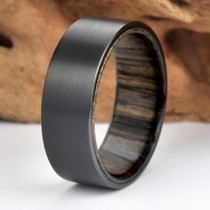 a wedding ring with black wood inlays on it, sitting next to a piece of driftwood