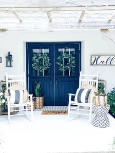 two white rocking chairs sitting in front of a blue door with wreaths on it