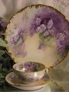 an antique tea cup and saucer with purple flowers on the rim, next to a plate