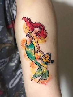 the little mermaid tattoo on the arm is painted with watercolors and has an orange fish