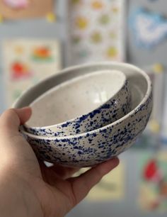 a hand holding two white and blue bowls in front of a wall with pictures on it