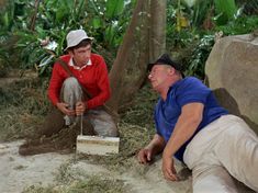 two men are sitting on the ground in front of some trees and plants, one is holding a box