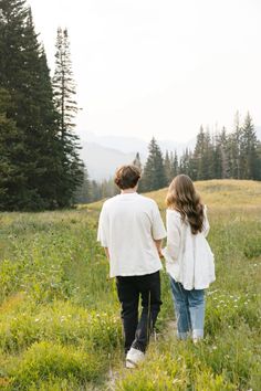 a man and woman are walking through the grass in front of pine trees on a hill